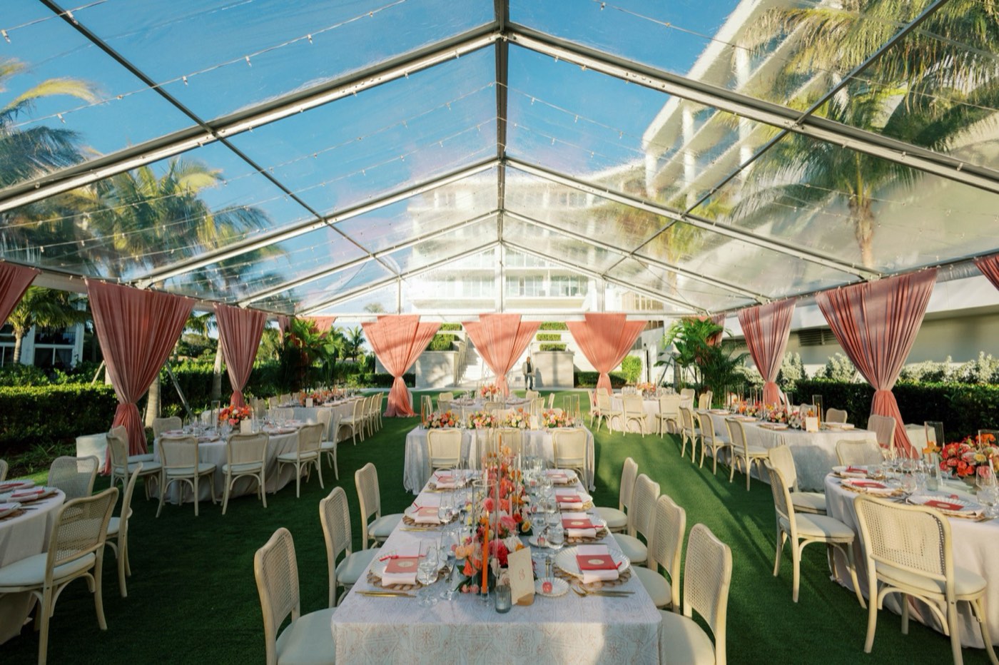 Clear tent reception with pink draping, palm trees, and arranged dining tables
